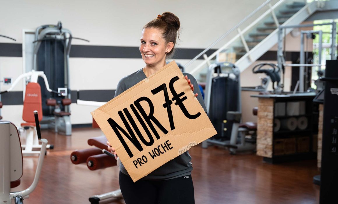 Simone vom TONUS SportsClub bewirbt die aktuelle Aktion mit einem Pappschild – dieses hat die Aufschrift „Nur 7 € pro Woche“. Im Hintergrund sieht man die Trainingsfläche.